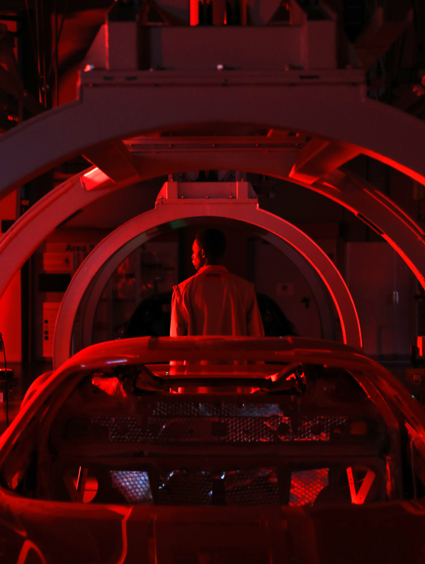 A man seen from behind standing in front of a vehicle under construction inside Maranello Car Factory in a red light