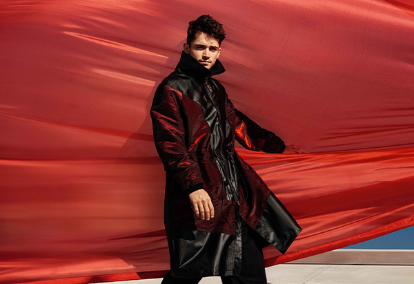 Half-length picture of Charles Leclerc wearing an iridescent burgundy and black jacket in front of a red drape blown by the wind 