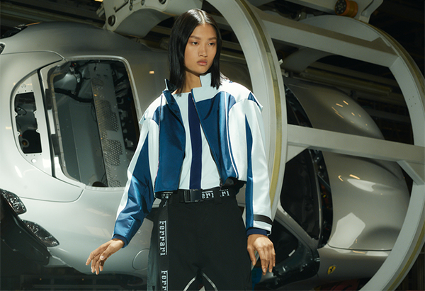 Half-length picture of a woman wearing white and blue blouson in front of a car inside Ferrari factory