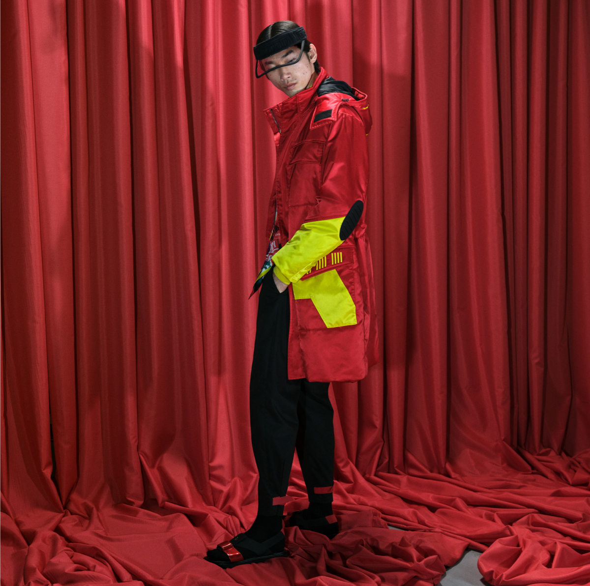Model in front of red curtains wearing black visor and red parka